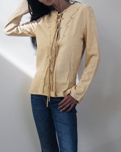 A woman wearing a light beige long-sleeve blouse with decorative tie-front laces and dark blue jeans, standing against a plain light gray wall.