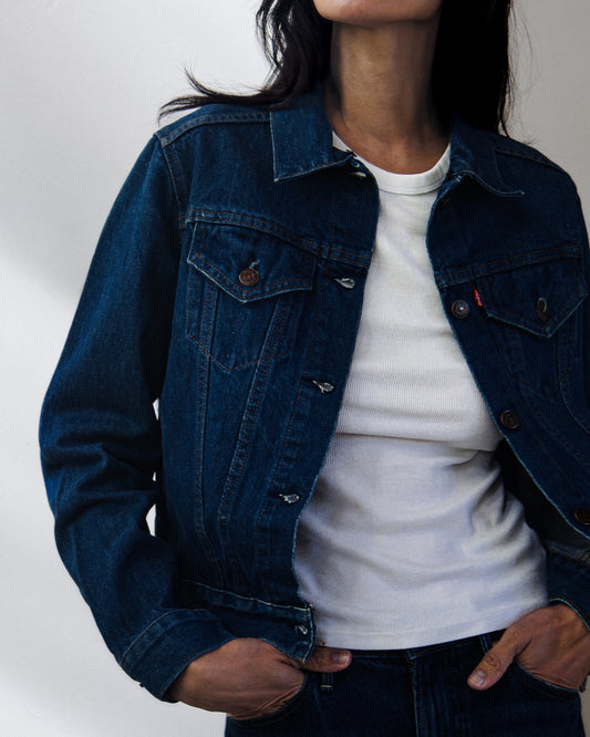 A close-up front view of a woman wearing a dark indigo denim jacket, showing the back pockets and buttoned hem against a plain light gray wall.