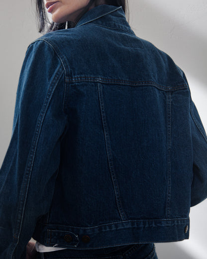 A close-up back view of a woman wearing a dark indigo denim jacket, showing the back pockets and buttoned hem against a plain light gray wall.