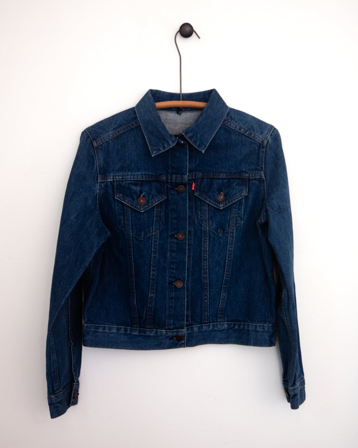 A close-up front view of a dark indigo denim jacket, showing the back pockets and buttoned hem hanging against a plain light gray wall.