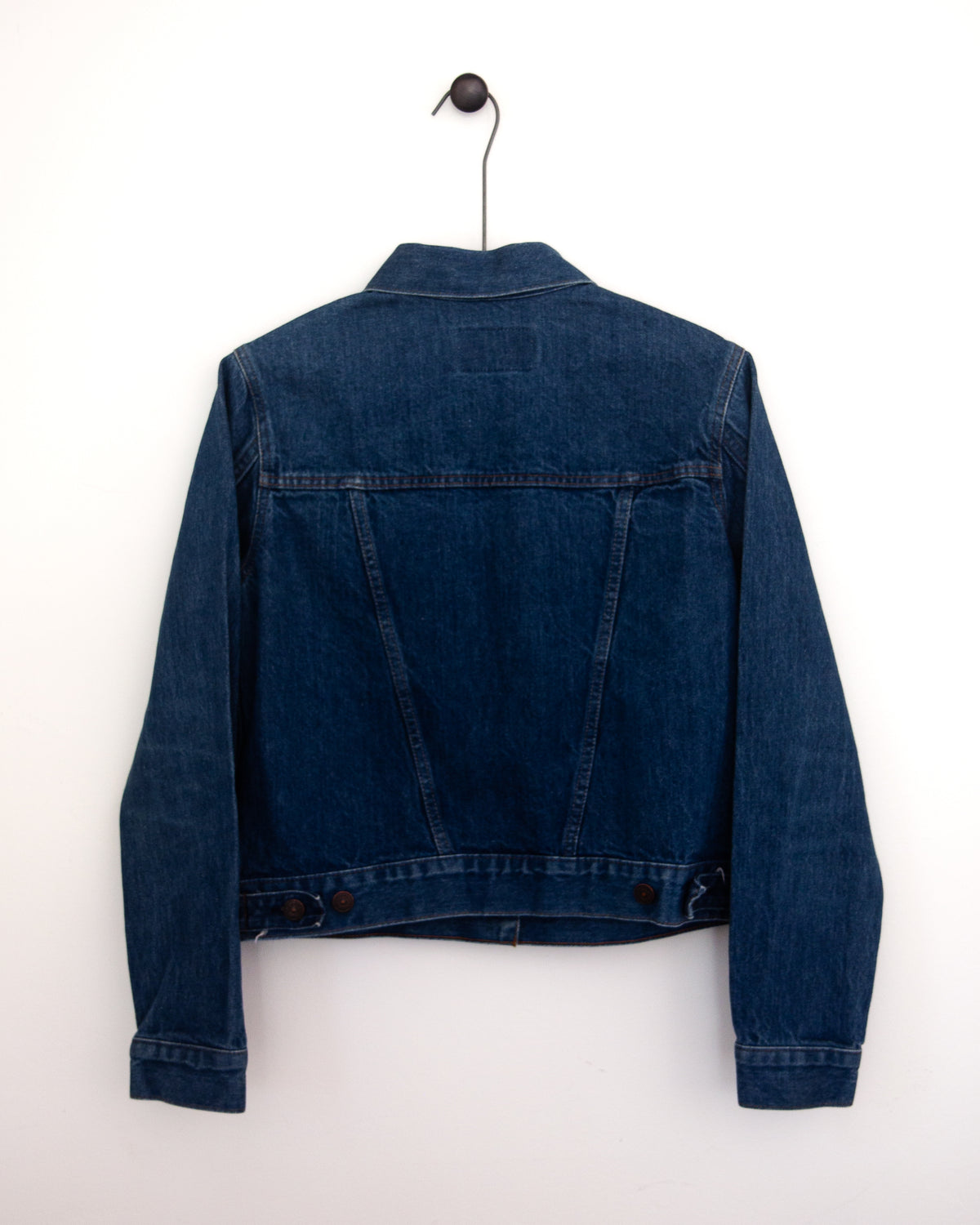 A close-up back view of a dark indigo denim jacket, showing the back pockets and buttoned hem hanging against a plain light gray wall.