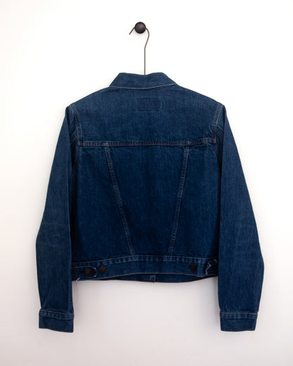 A close-up back view of a dark indigo denim jacket, showing the back pockets and buttoned hem hanging against a plain light gray wall.