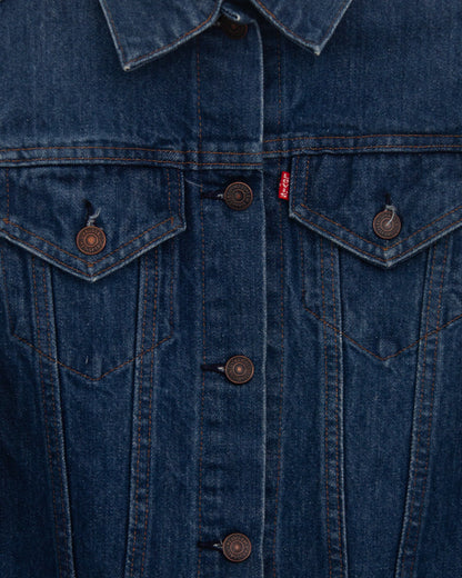 A close-up back view of a dark indigo denim jacket, showing the back pockets and buttoned hem hanging against a plain light gray wall.