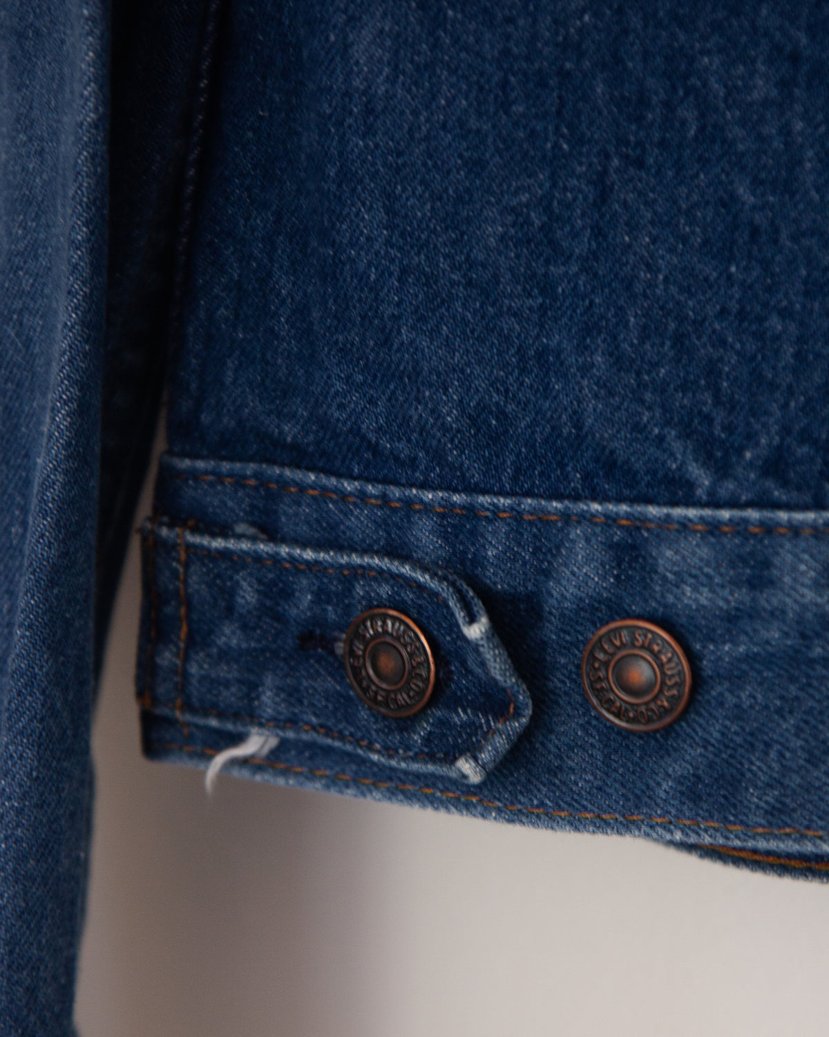 Close-up of a dark blue denim jacket showing the cuff detail with two bronze-colored metal buttons and contrast stitching.