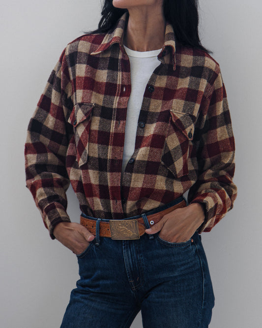 A person wearing a long-sleeve plaid button-up shirt in burgundy, tan, and black over a white t-shirt, paired with blue jeans and a brown leather belt, against a plain light gray background.