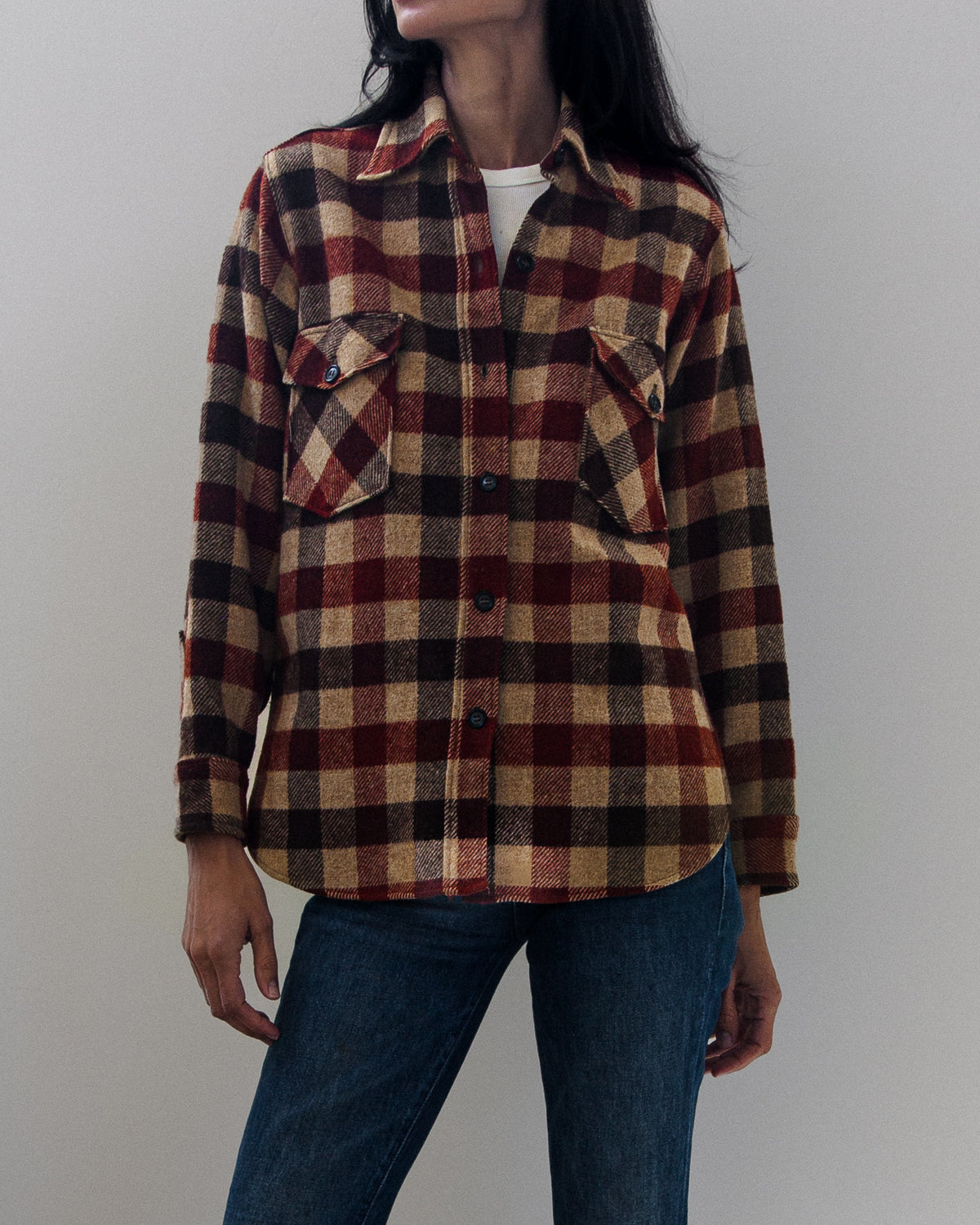 A person wearing a long-sleeve plaid button-up shirt in burgundy, tan, and black over a white t-shirt, paired with blue jeans against a plain light gray background.