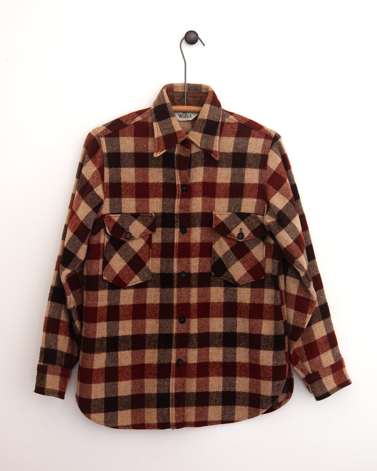 Close-up of a red, beige, and black plaid flannel shirt jacket with a chest pocket and button closure on a hanger against a white wall.