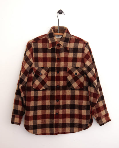 Close-up of a red, beige, and black plaid flannel shirt jacket with a chest pocket and button closure on a hanger against a white wall.