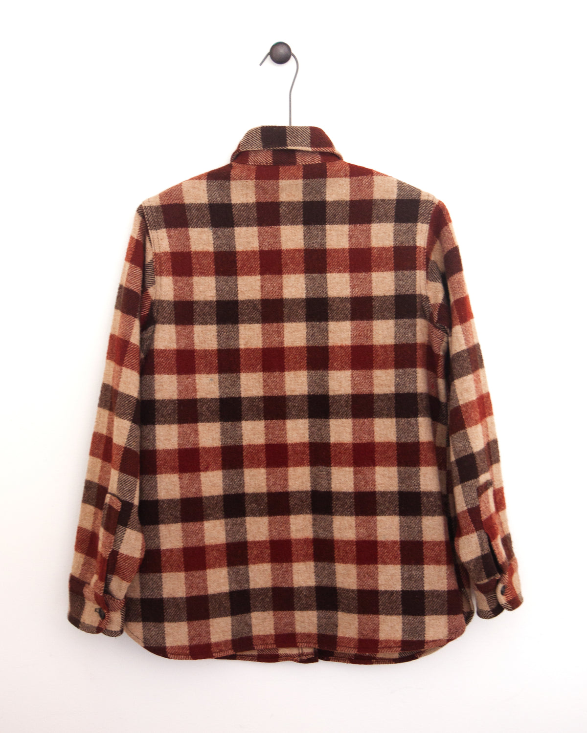Close-up back view of a red, beige, and black plaid flannel shirt jacket with a chest pocket and button closure on a hanger against a white wall.