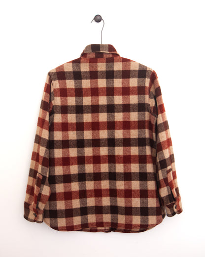 Close-up back view of a red, beige, and black plaid flannel shirt jacket with a chest pocket and button closure on a hanger against a white wall.