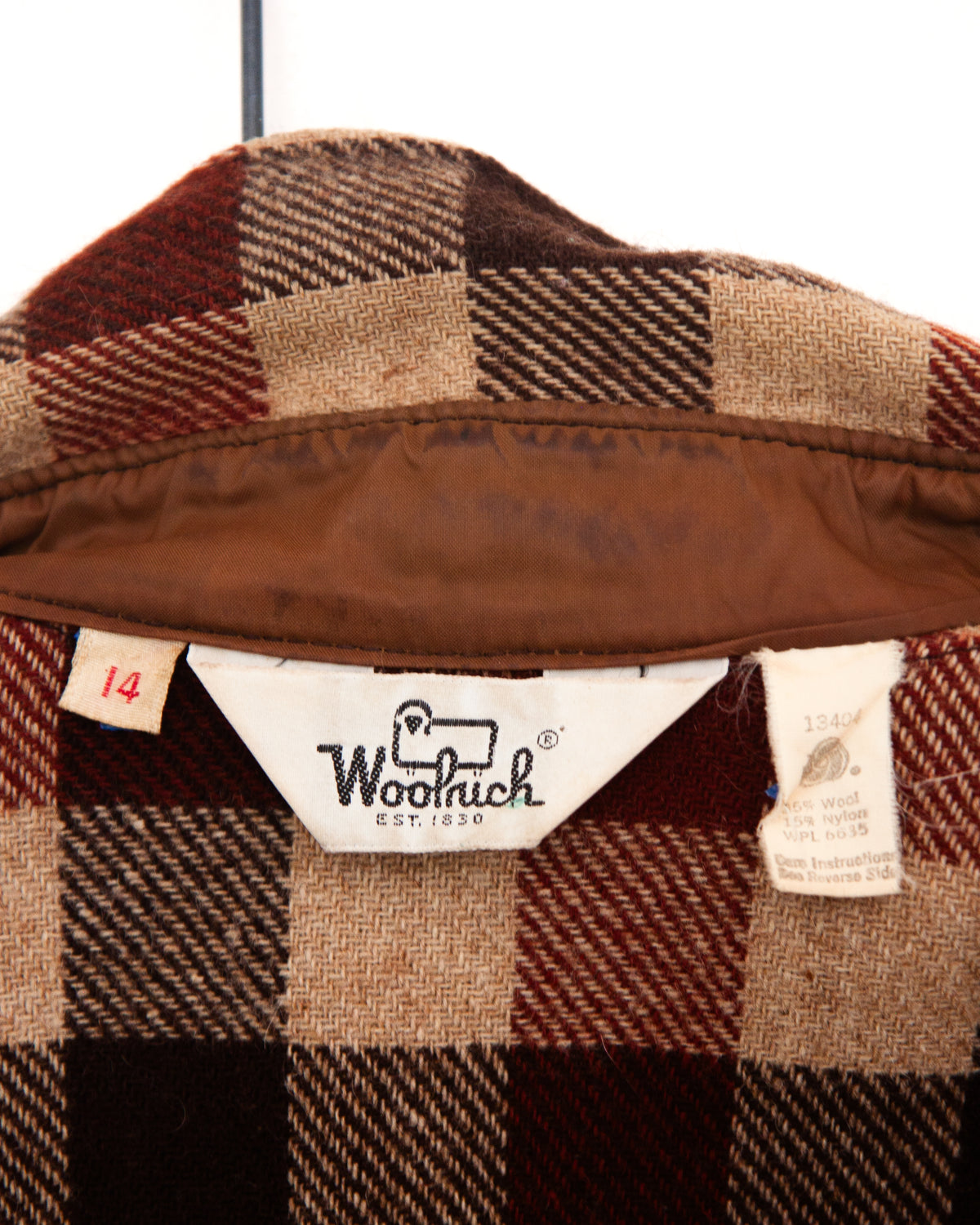 A close-up of a plaid wool or flannel fabric sleeve, showing a checkered pattern in dark red, tan, brown, and black with a Woolrich tags on a plain white background.