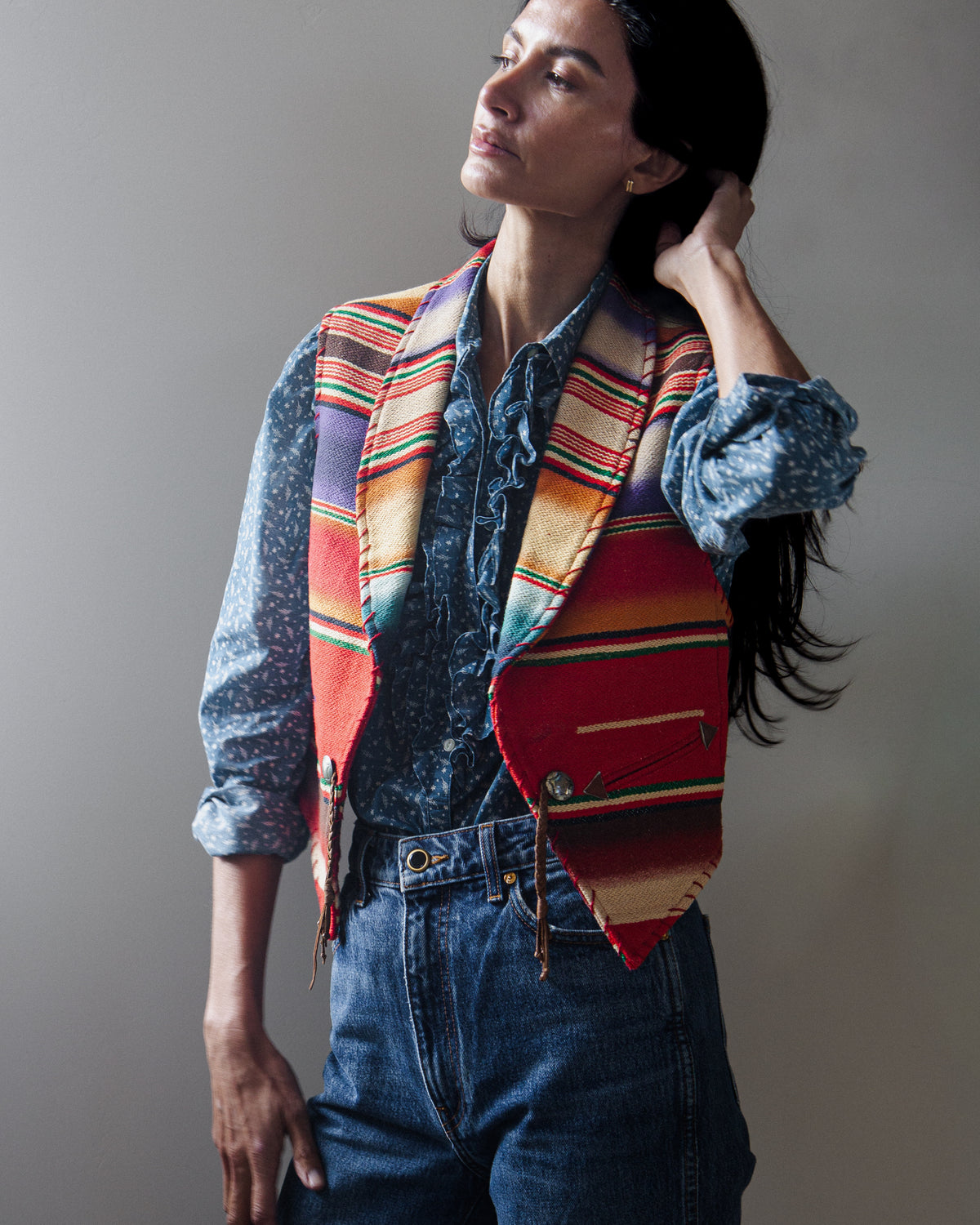 A woman wearing a colorful striped vest with fringe over a blue patterned shirt and blue jeans, posed against a neutral gray studio background.