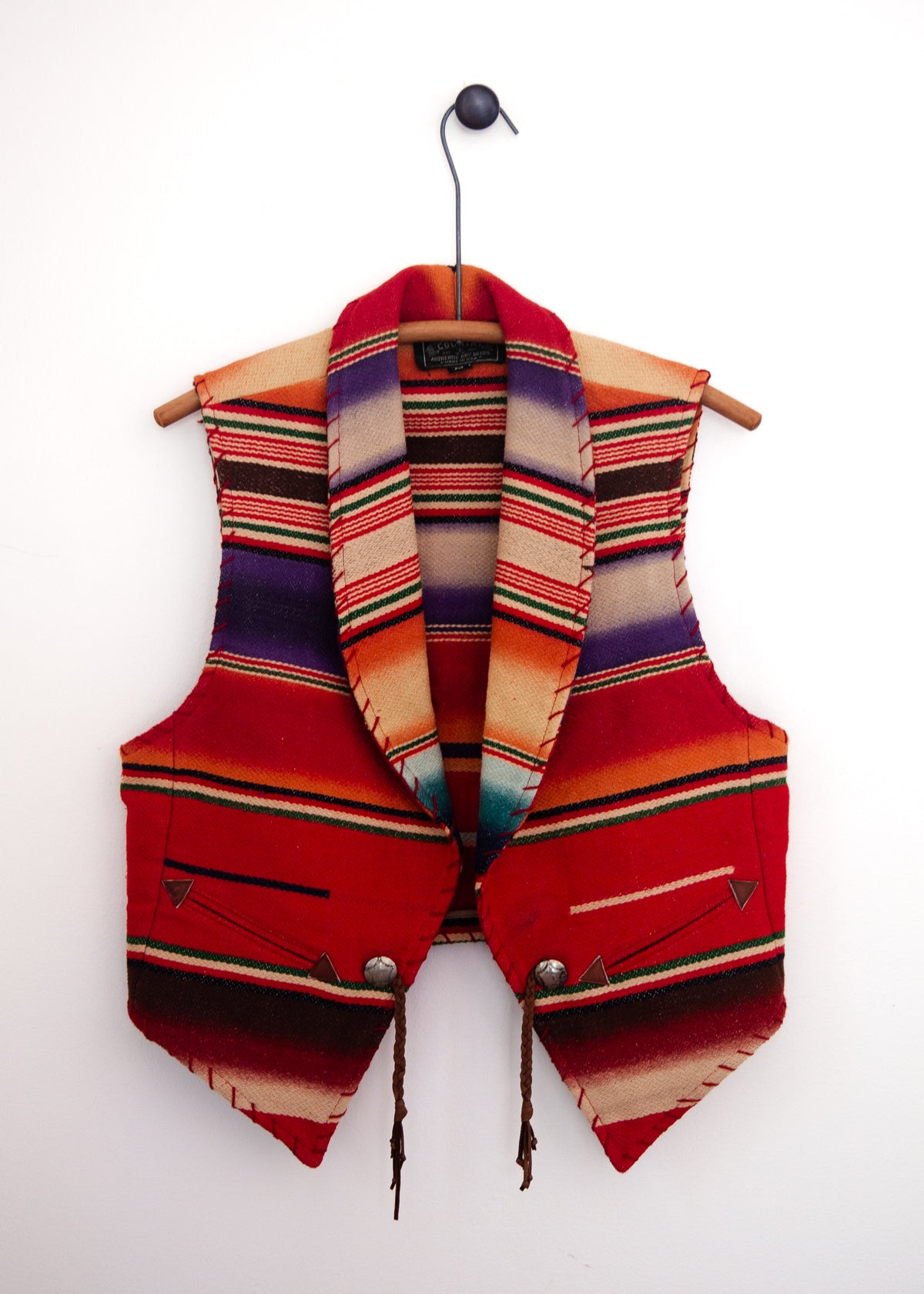 A sleeveless woven vest with a colorful serape-style striped pattern, hanging on a wooden hanger against a plain white wall.