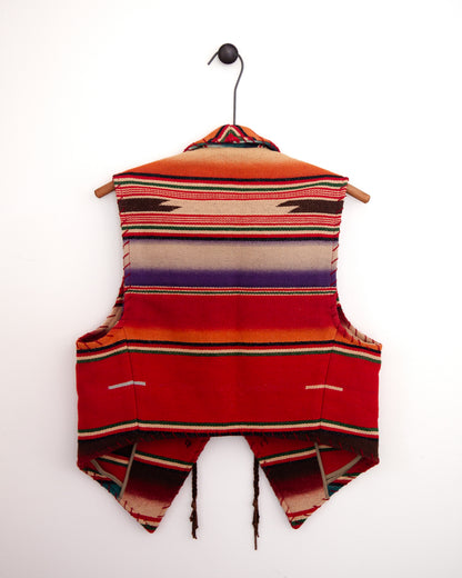 A sleeveless woven vest with a striped serape pattern in red, orange, beige, purple, and black, hanging on a wooden hanger against a plain white wall.