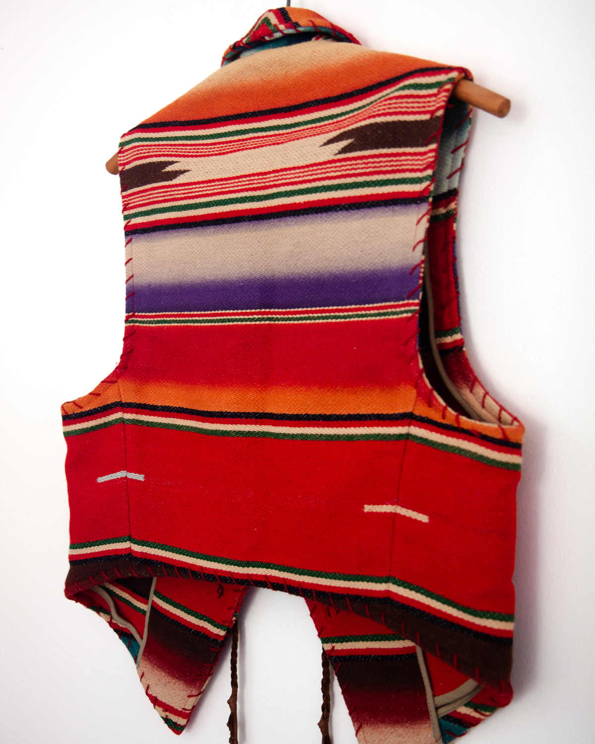 A sleeveless woven vest with a colorful serape-style striped pattern, hanging on a wooden hanger against a plain white wall.