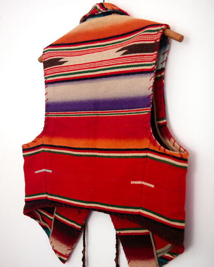 A sleeveless woven vest with a colorful serape-style striped pattern, hanging on a wooden hanger against a plain white wall.
