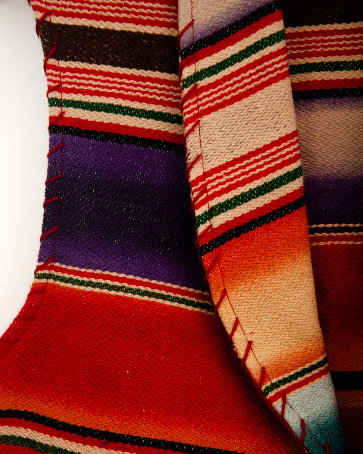 Detail of a sleeveless woven vest with a colorful serape-style striped pattern, hanging on a wooden hanger against a plain white wall.