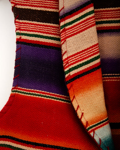 Detail of a sleeveless woven vest with a colorful serape-style striped pattern, hanging on a wooden hanger against a plain white wall.