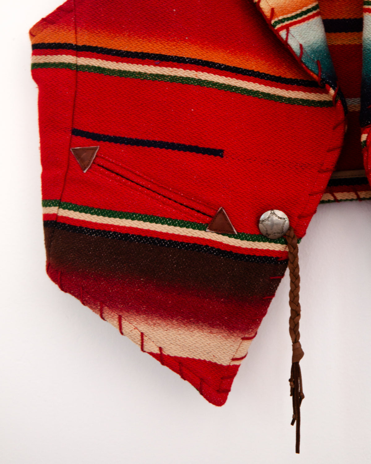 A close-up of a folded red woven textile with horizontal stripes in green, cream, and dark brown, featuring a metal snap button and a braided cord on a plain white background.
