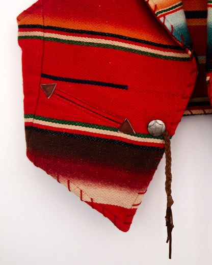 A close-up of a folded red woven textile with horizontal stripes in green, cream, and dark brown, featuring a metal snap button and a braided cord on a plain white background.