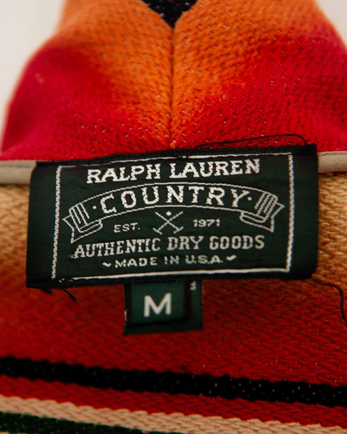 Detail of a sleeveless woven vest Ralph Lauren Country Authentic Dry Goods tag with a colorful serape-style striped pattern, hanging on a wooden hanger against a plain white wall.