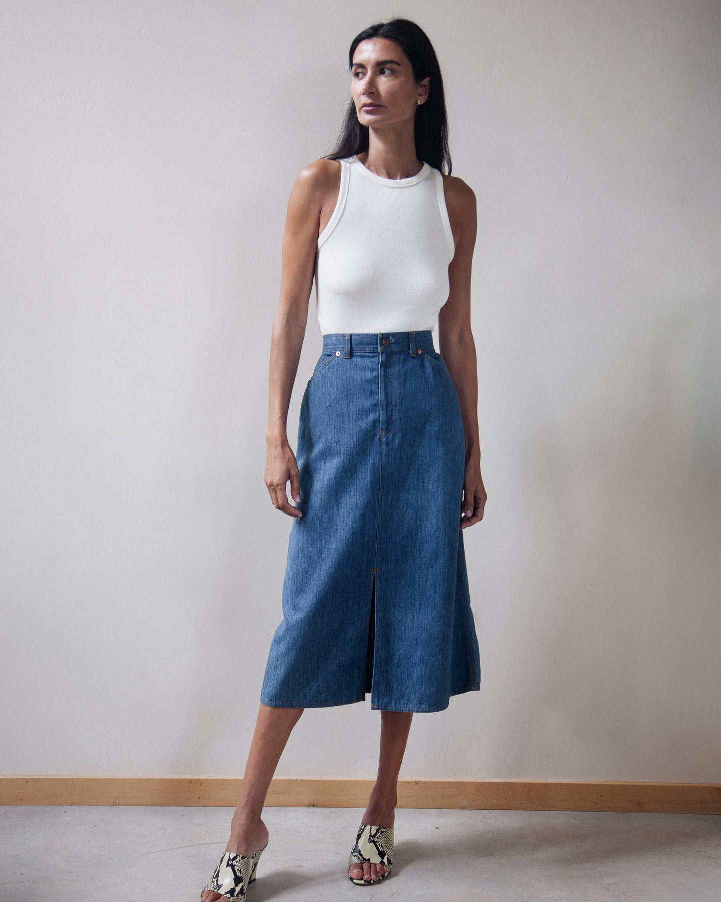 A model wearing a blue denim midi skirt with a front slit, paired with a white ribbed tank top and patterned sandals, standing against a neutral off-white wall.