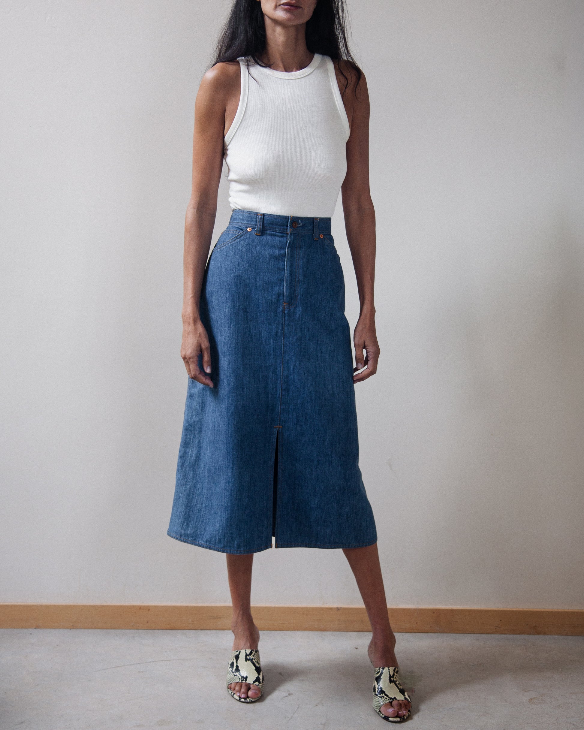 A model wearing a blue denim midi skirt with a front slit, paired with a white ribbed tank top and patterned sandals, standing against a neutral off-white wall.