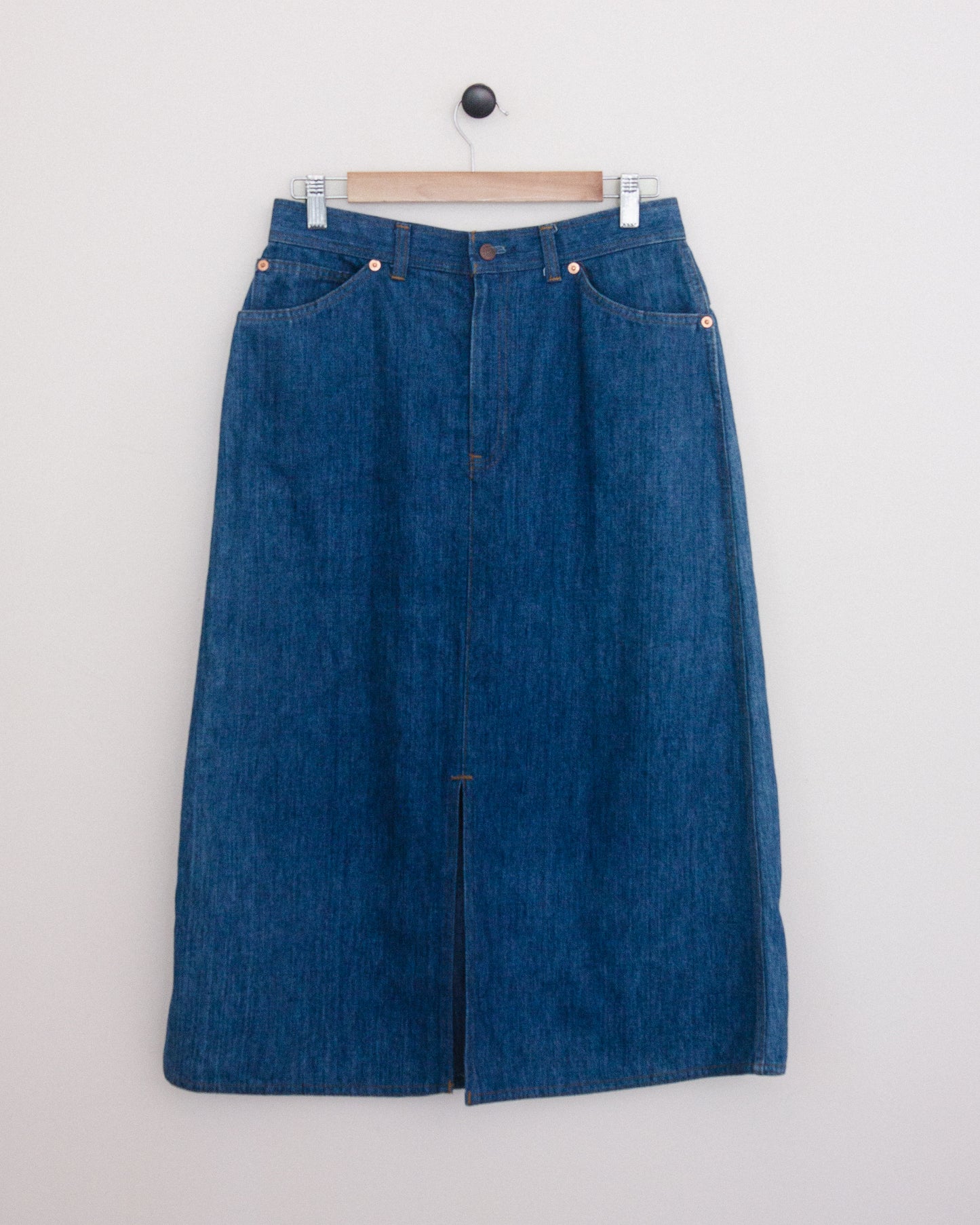 A blue denim midi skirt with a front slit, hanging on a wooden hanger against a plain white wall.