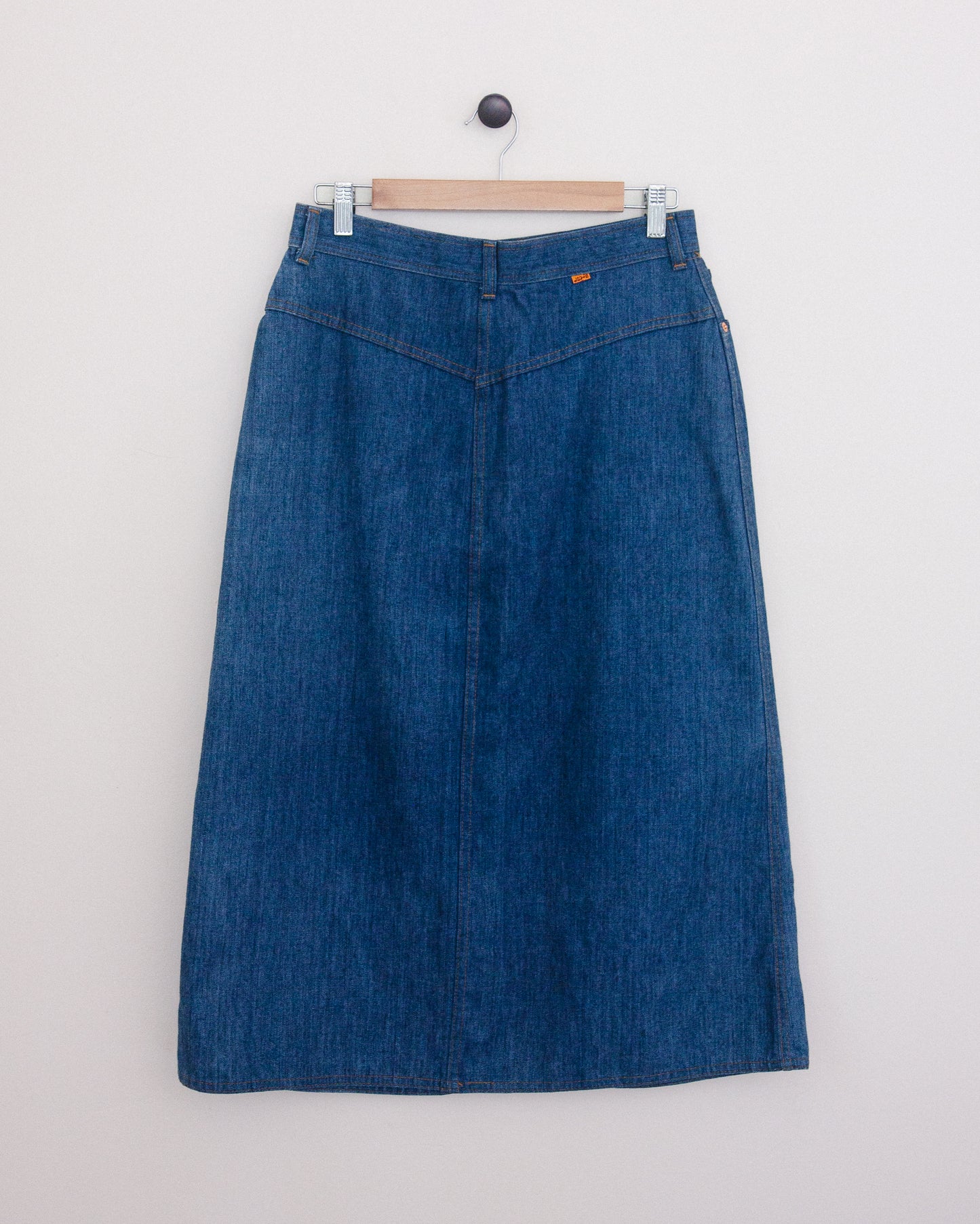 Back of a blue denim midi skirt hanging on a wooden hanger against a plain white wall.
