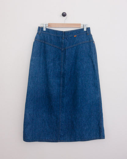 Back of a blue denim midi skirt hanging on a wooden hanger against a plain white wall.