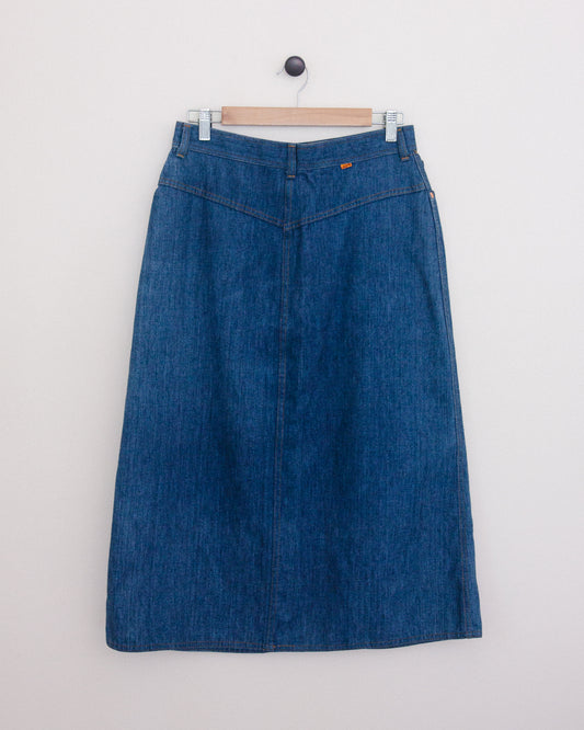 Back of a blue denim midi skirt hanging on a wooden hanger against a plain white wall.