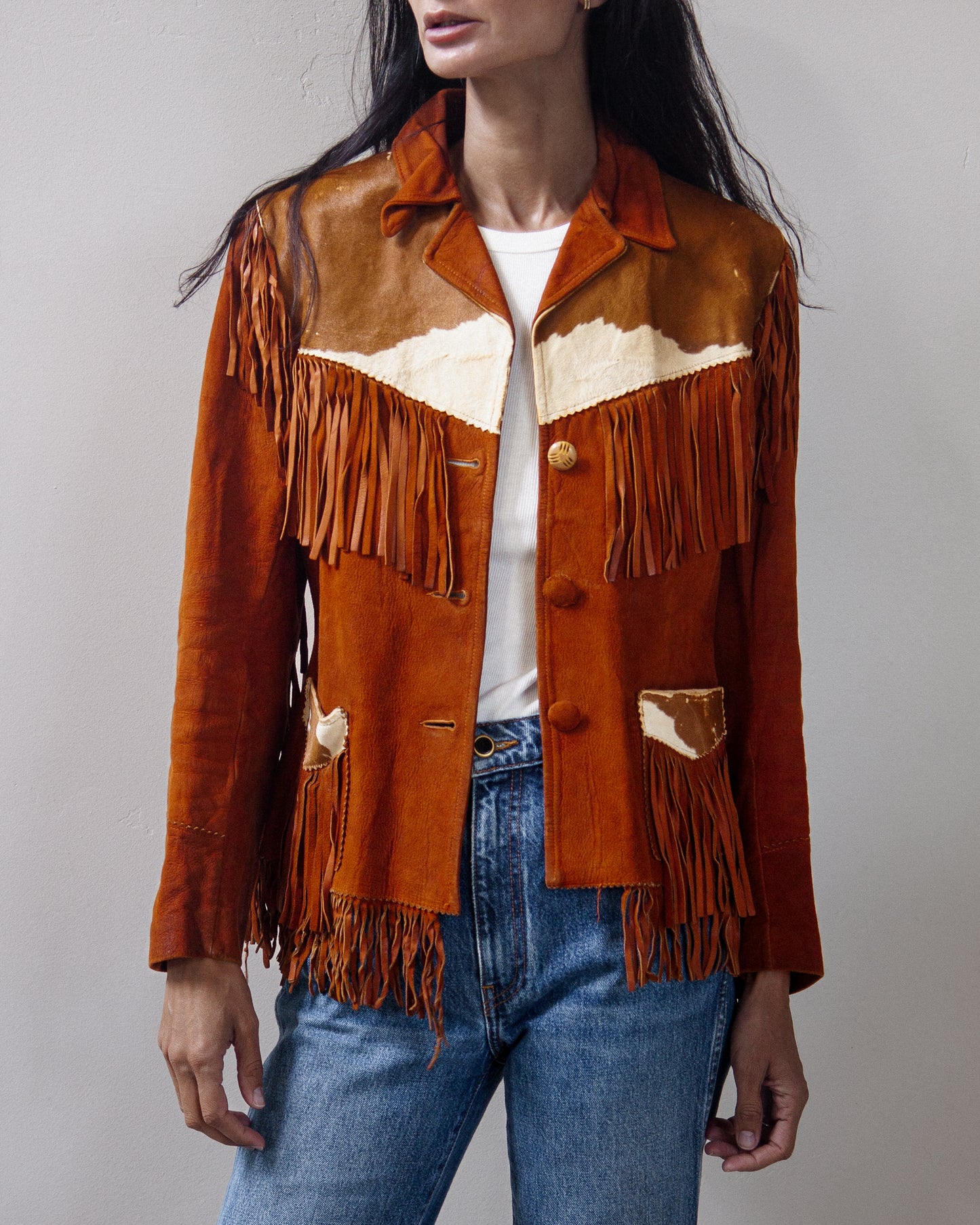 A woman wearing a brown suede fringe jacket with cream-colored panels and blue jeans, photographed against a neutral gray studio background.