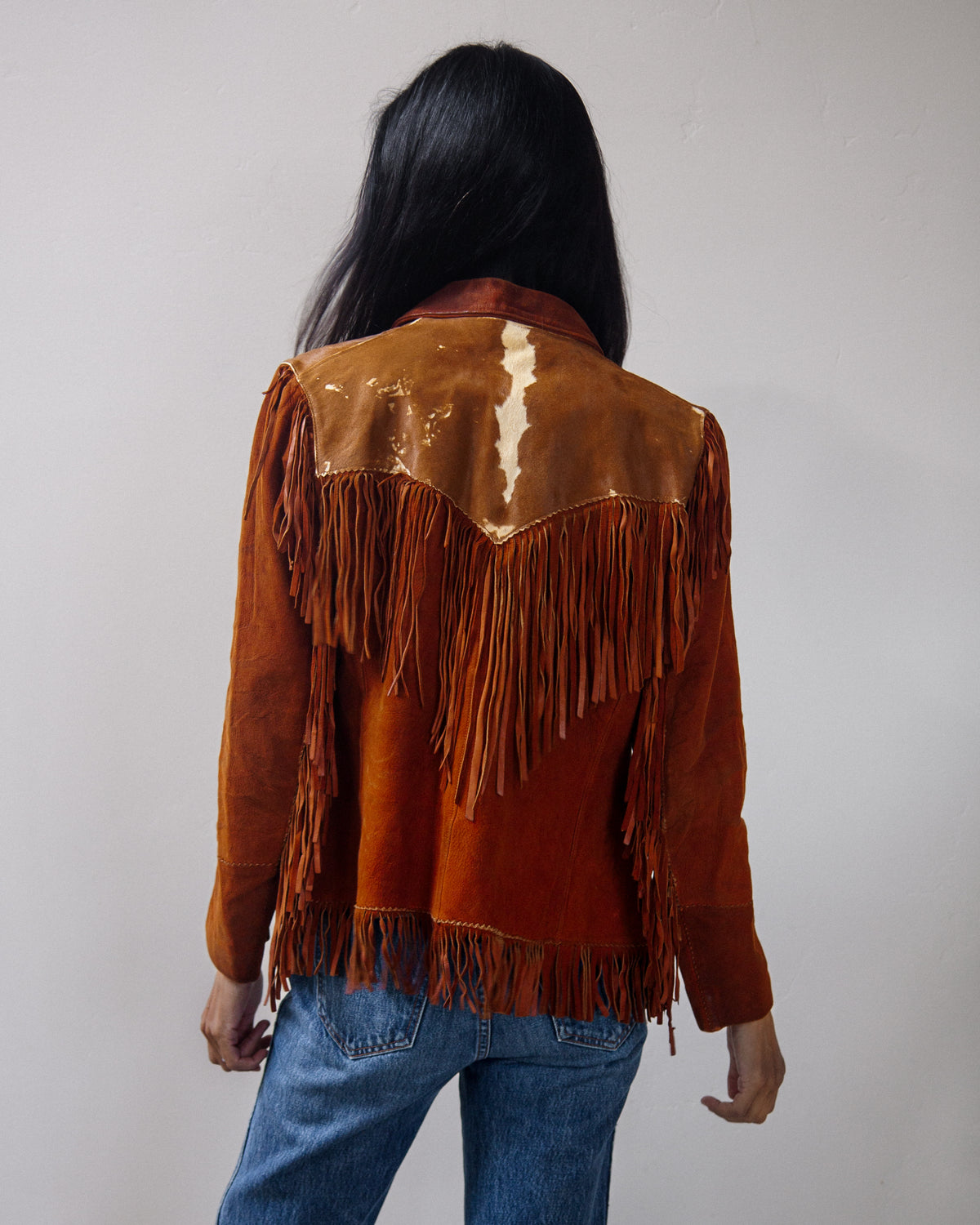A person shown from the back wearing a brown suede fringe jacket with a cow-print shoulder panel and blue jeans, standing against a plain light gray wall.