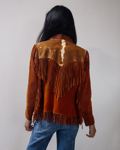 A person shown from the back wearing a brown suede fringe jacket with a cow-print shoulder panel and blue jeans, standing against a plain light gray wall.