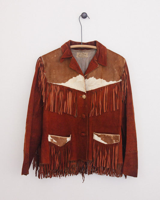 A vintage brown suede fringe jacket with a western design, featuring a white cowhide patch on the chest and matching patches on the pockets, hanging on a wooden hanger against a white wall.