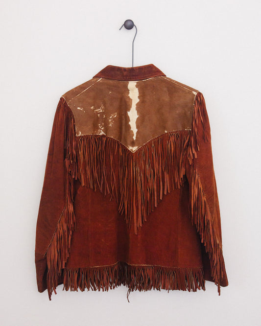 Back of a brown suede fringe jacket with contrasting tan leather shoulder panels and a central cream-colored vertical stripe, hanging on a black wire hanger against a plain white wall.