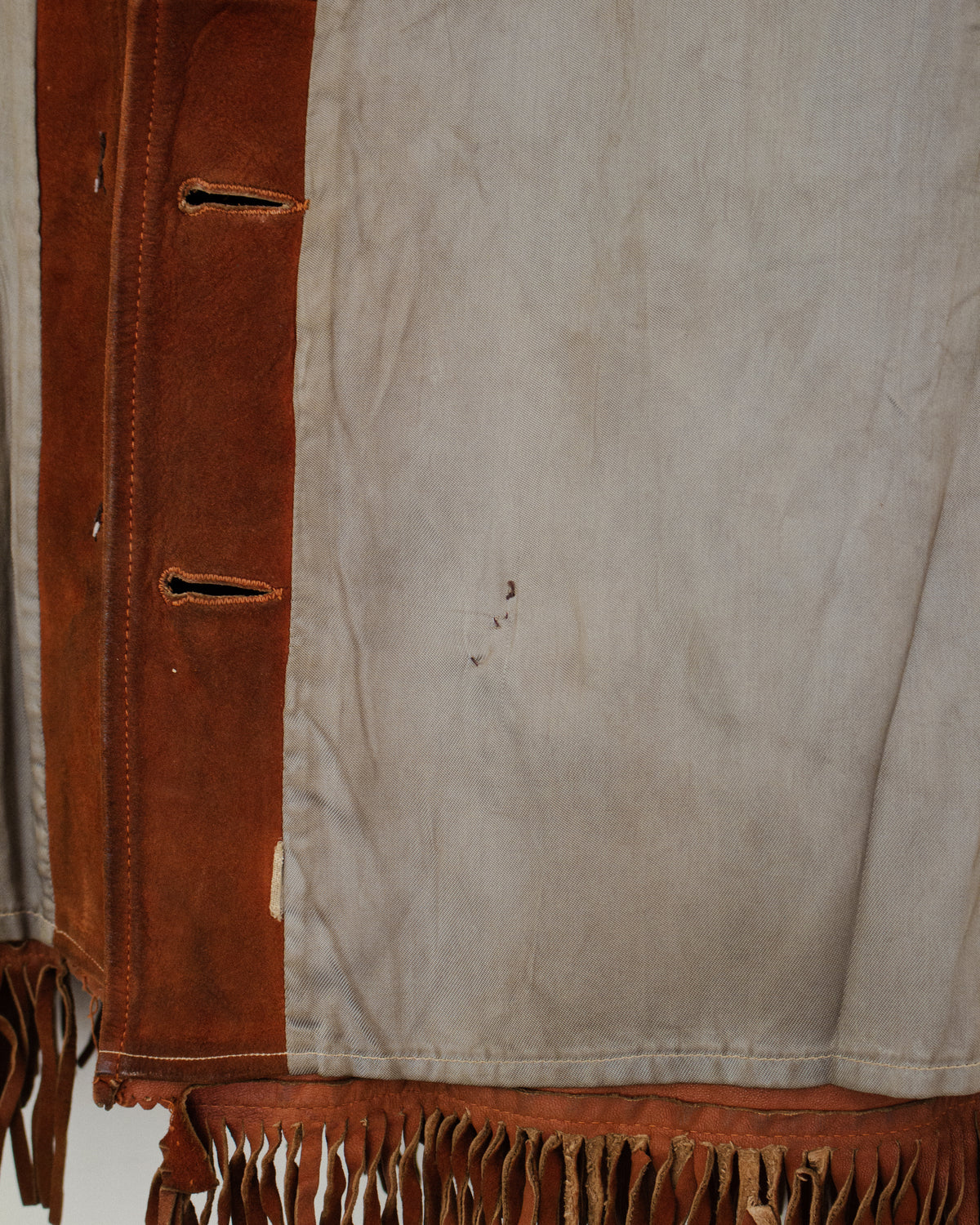 Close-up of a fringed garment or textile with a light beige suede-like body and a vertical brown suede panel with stitched openings and fringe. along the bottom.