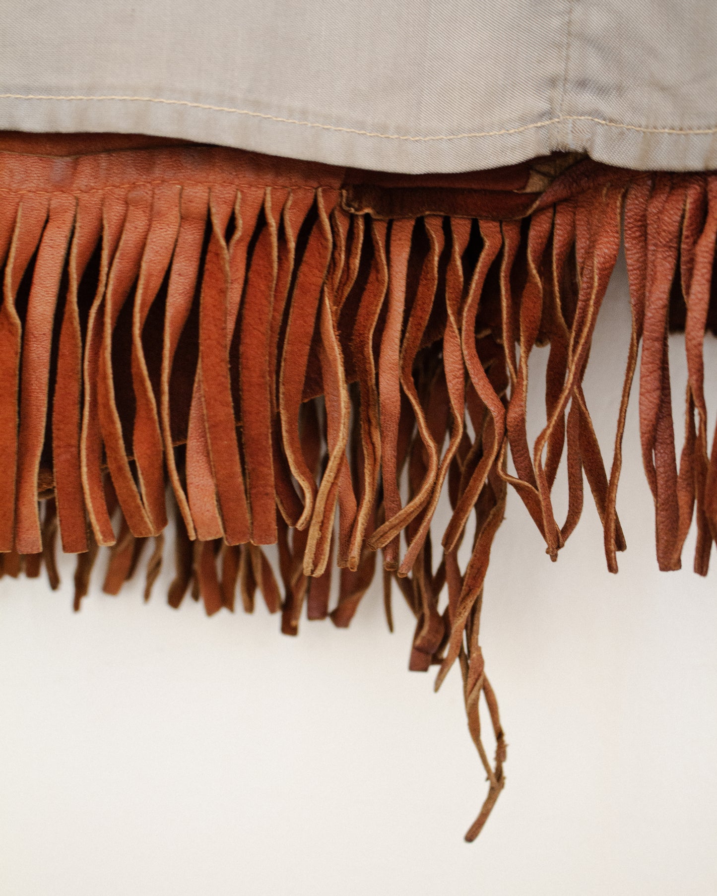Close-up of a brown leather fringe trim hanging from a light beige interior lining of fabric against a plain white background.