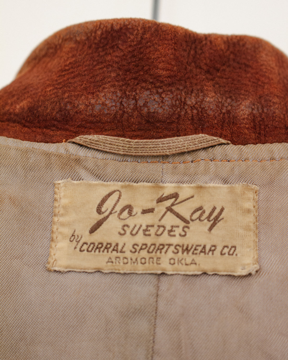Close-up of the interior neck label of a vintage jacket, showing a beige fabric patch with brown text that reads 'Jo-Kay SUEDES by CORRAL SPORTSWEAR CO. ARDMORE OKLA.