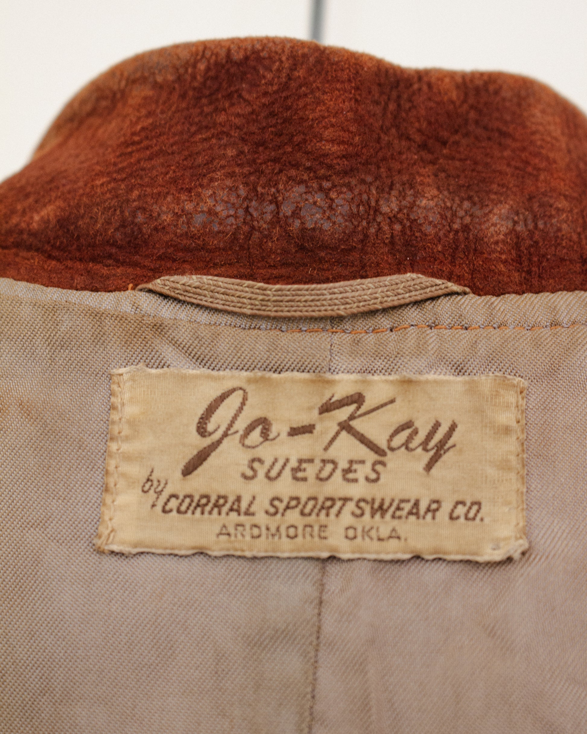 Close-up of the interior neck label of a vintage jacket, showing a beige fabric patch with brown text that reads 'Jo-Kay SUEDES by CORRAL SPORTSWEAR CO. ARDMORE OKLA.