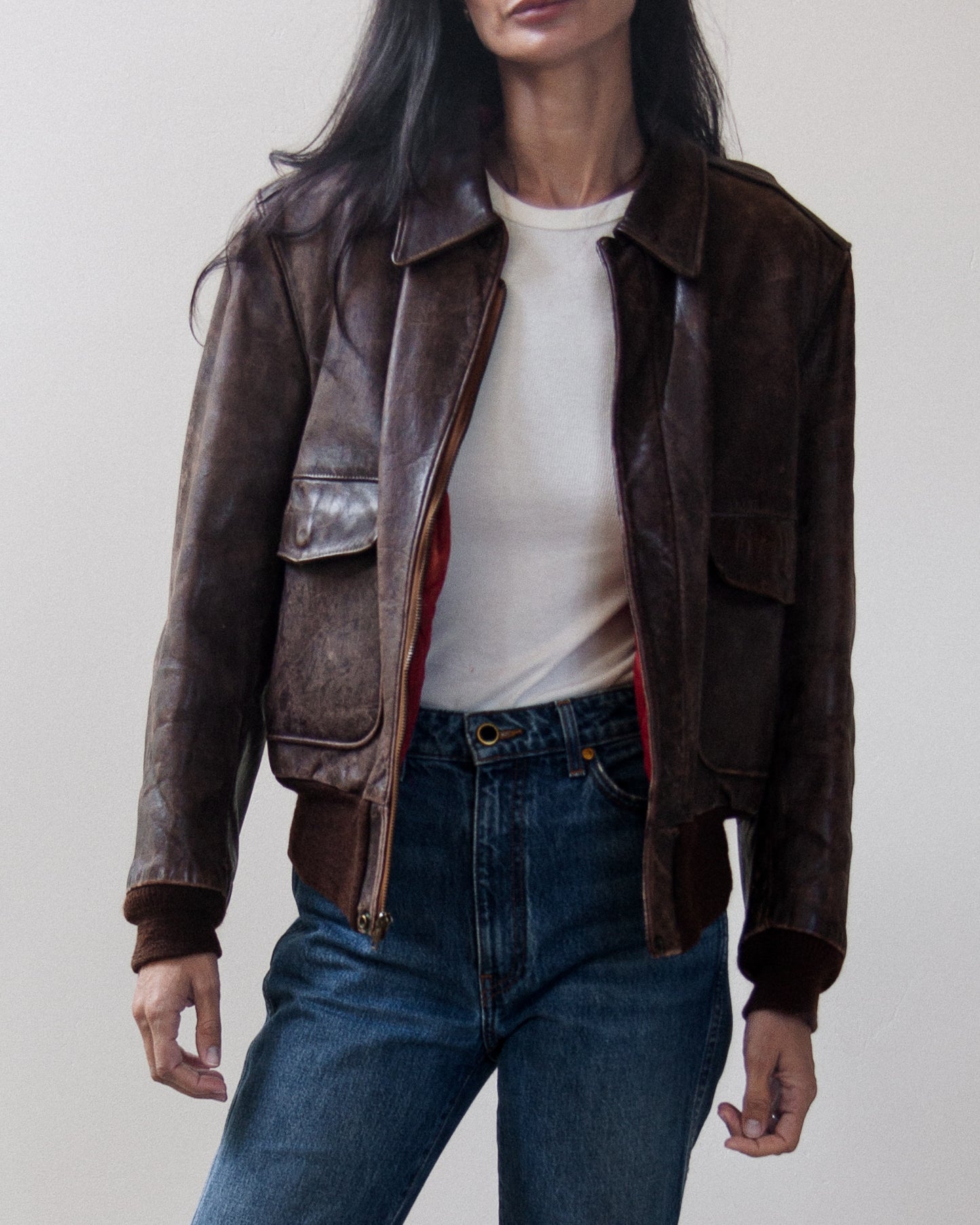 A woman wearing a dark brown distressed leather aviator-style jacket over a cream t-shirt and blue jeans, shown from the neck down against a plain light gray background.