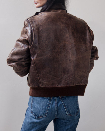 A person shown from the back wearing a distressed dark brown leather jacket with ribbed collar, cuffs, and hem, paired with blue jeans against a plain light gray background.