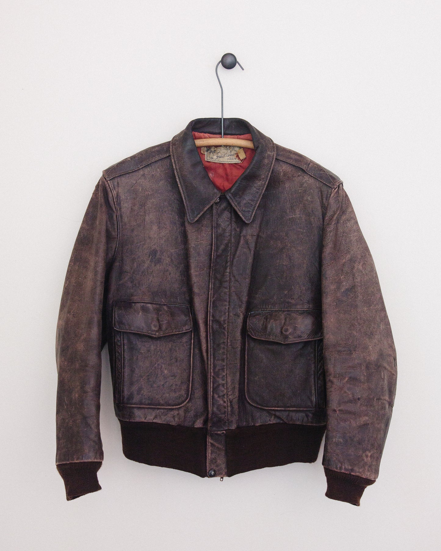 A distressed dark brown leather jacket hanging on a black hanger against a plain white wall. The jacket features a faded brown finish, a pointed collar, a front zipper, and two large front flap pockets.