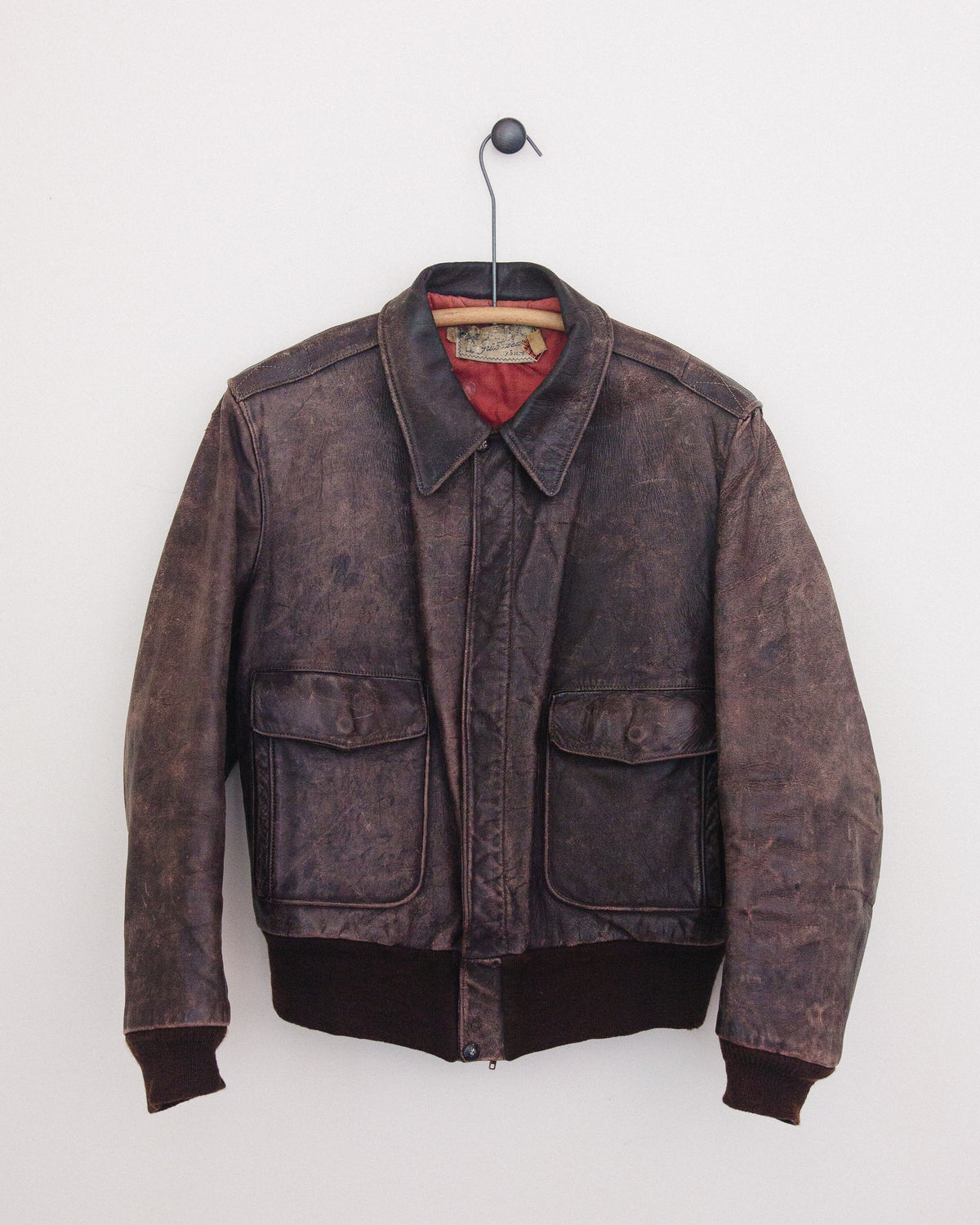 A distressed dark brown leather jacket hanging on a black hanger against a plain white wall. The jacket features a faded brown finish, a pointed collar, a front zipper, and two large front flap pockets.