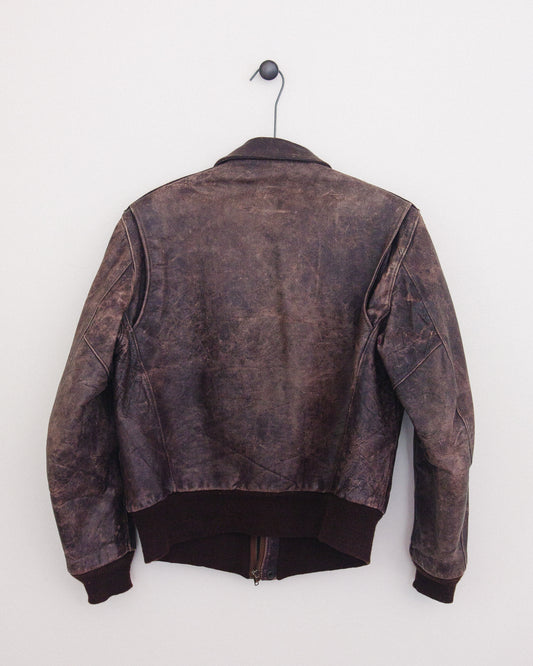 A dark brown distressed leather jacket hanging on a black hanger against a plain white wall, showing the back view.