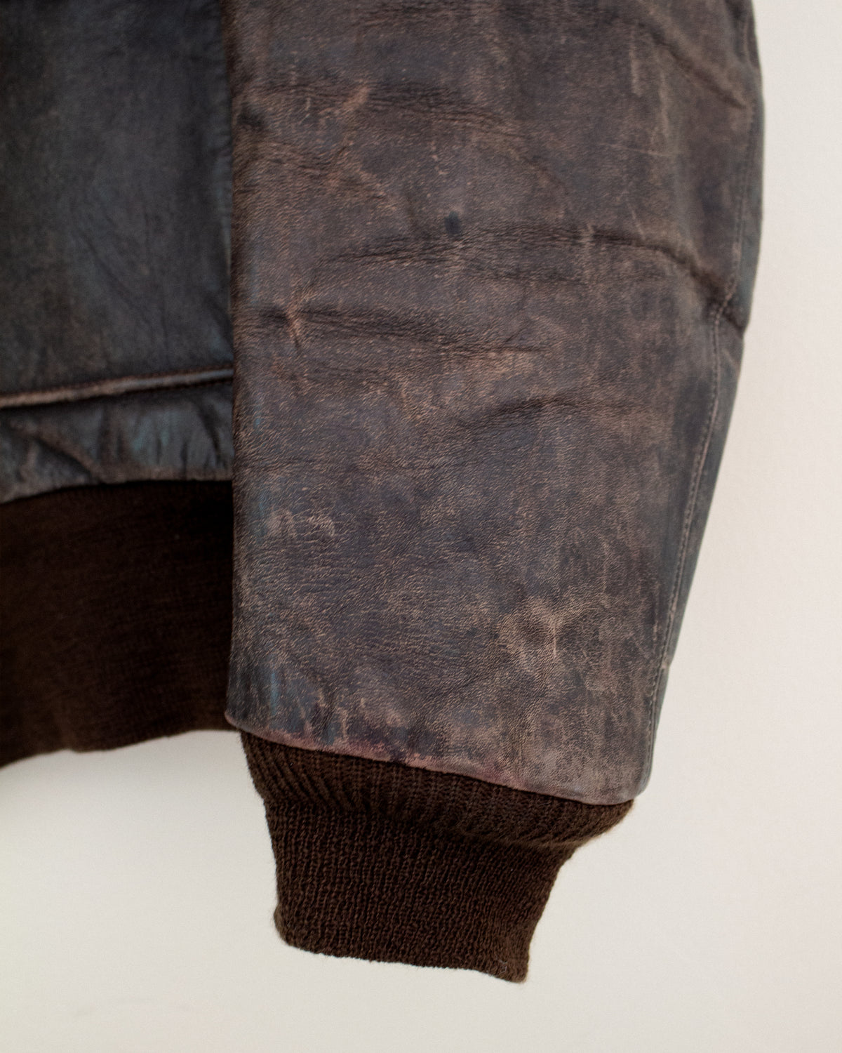 Close-up of a distressed dark brown leather jacket sleeve with a ribbed brown knit cuff, showing the textured leather and inner lining against a plain light background.
