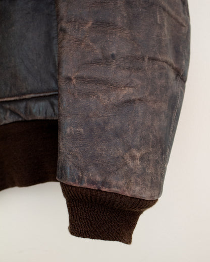 Close-up of a distressed dark brown leather jacket sleeve with a ribbed brown knit cuff, showing the textured leather and inner lining against a plain light background.