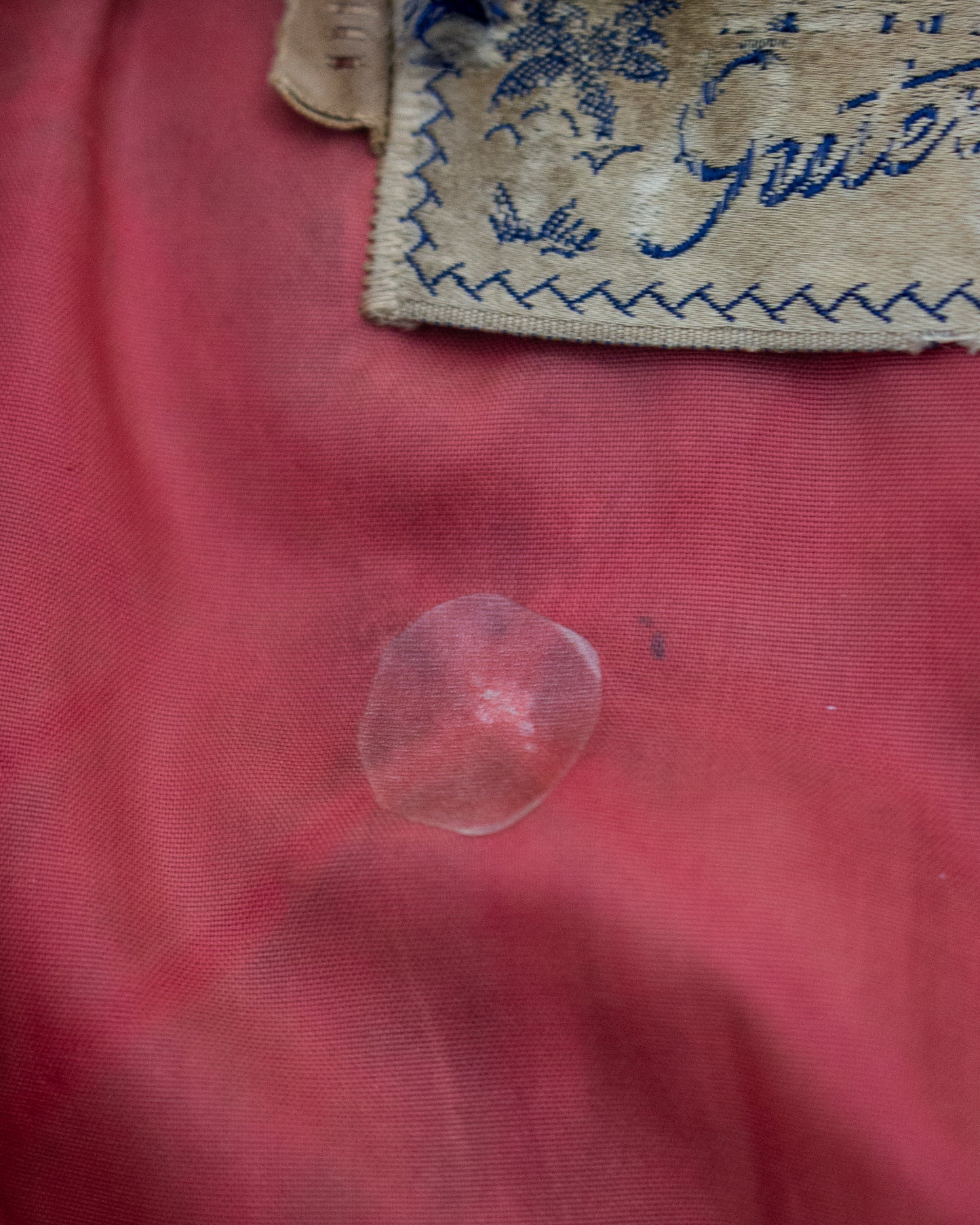 A close-up of a translucent stain on textured pink fabric, with a partially visible beige fabric tag showing blue embroidery at the top.