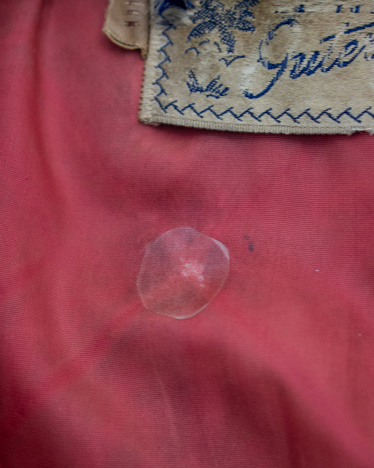 A close-up of a translucent stain on textured pink fabric, with a partially visible beige fabric tag showing blue embroidery at the top.