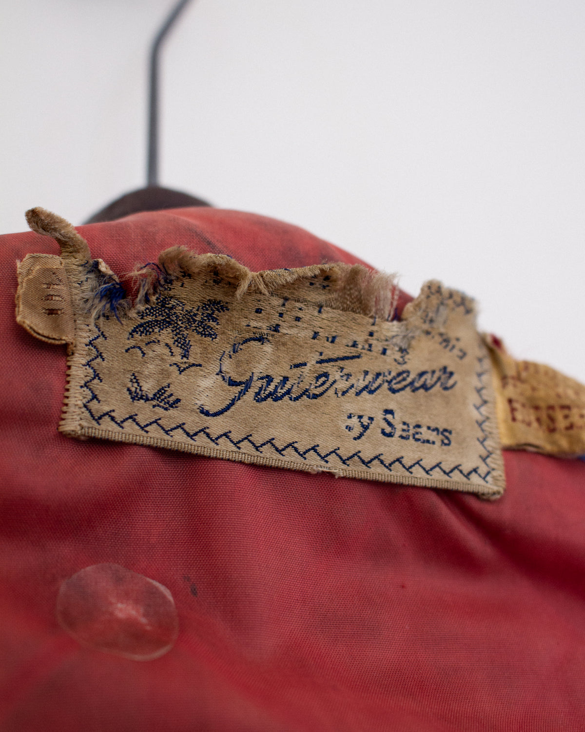 Close-up of a vintage red garment with a worn beige fabric label stitched in blue thread, photographed against a light background.